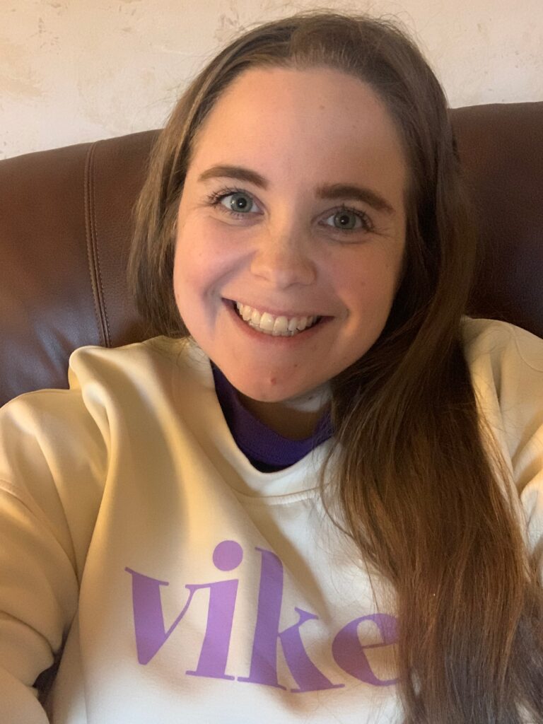 Emily Munns smiling at the camera while wearing a cream and yellow VIKES sweatshirt